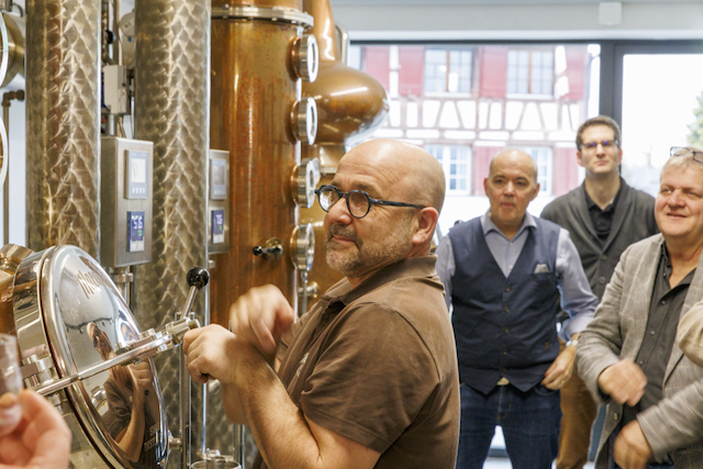 Im Vordergrund steht ein Mann mit Brille an einer Kupfer-Brennblase im Macardo Stil. Andere Teilnehmer des Macardo Brennkurses beobachten interessiert. Das Bild zeigt einen praktischen Teil des Macardo Brennkurses.